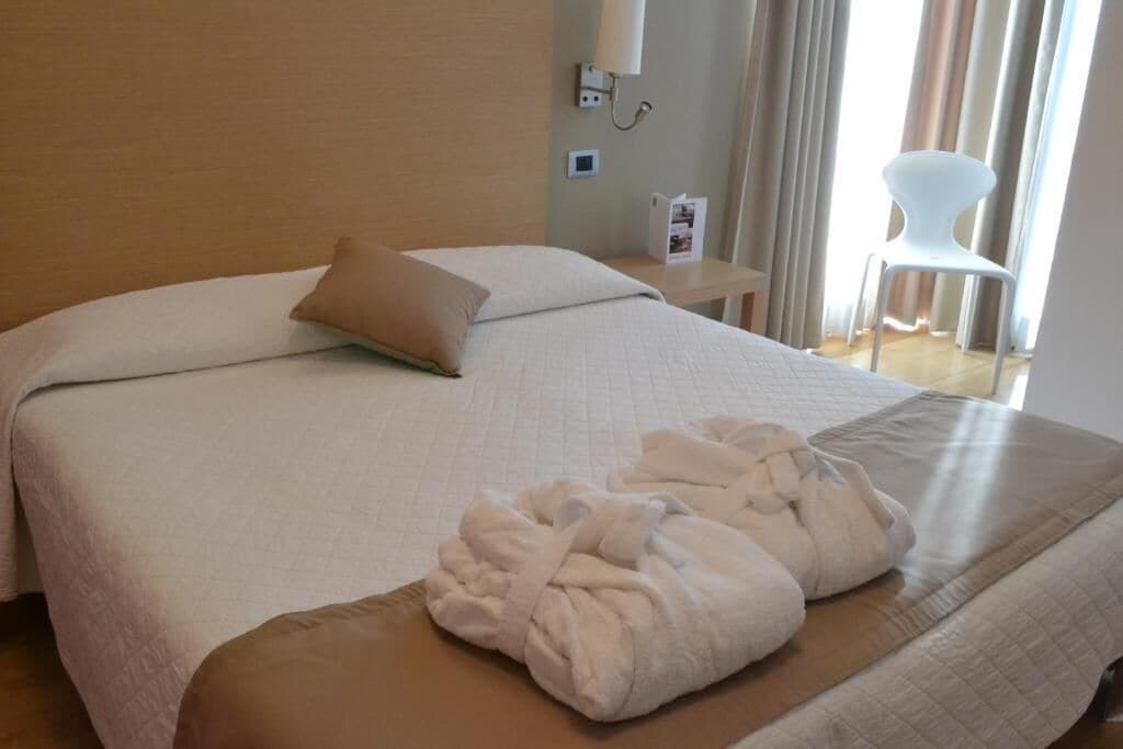 Ein Hotelzimmer mit Bett, Nachttisch mit Lampe, Stuhl und einem Fenster mit Vorhängen.