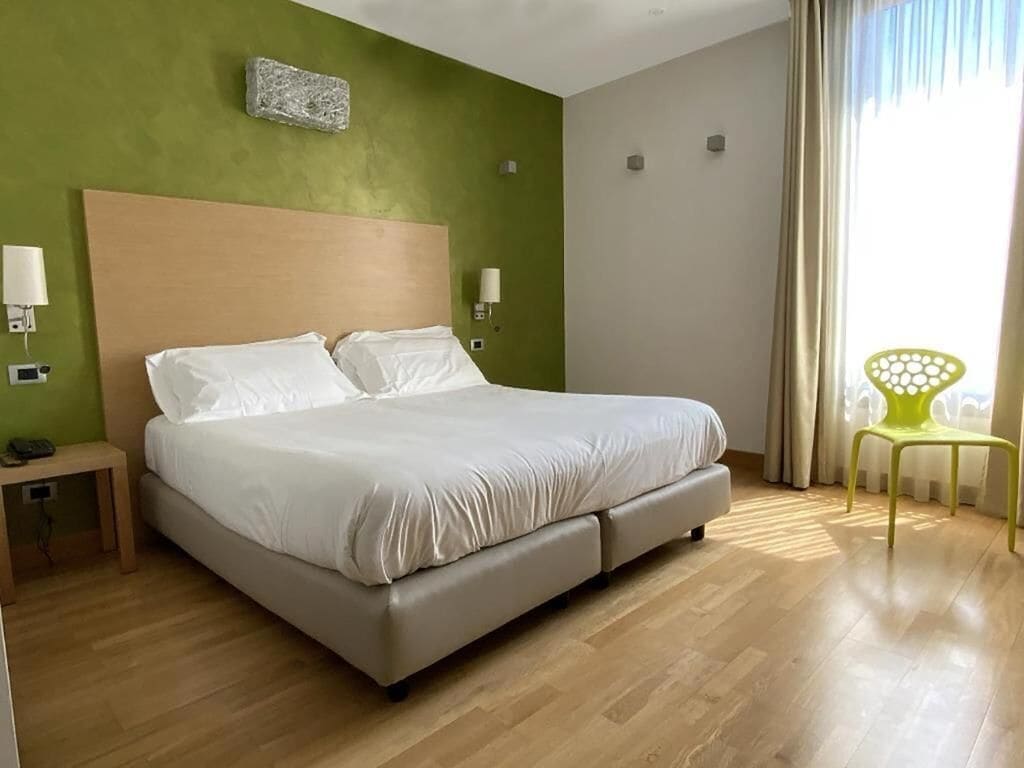 Habitación de hotel con cama, lámparas de noche, mesita de noche, una silla y ventana con cortinas.
