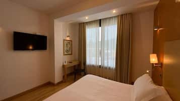 Quarto de hotel com cama, escrivaninha, cadeira, televisão e janela com cortinas.