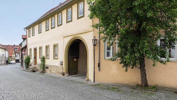 A building with a cobblestone street, a large arched doorway, and a tree in front. - Apartment 'Stadthaus' with Private Terrace, Balcony and Wi-Fi (Mellrichstadt)
