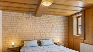 A bedroom with a brick wall, wooden ceiling beams, two bedside tables with lamps, a double bed with a plaid bedspread, and a window with a view. - Apartment 'Stadthaus' with Private Terrace, Balcony and Wi-Fi (Mellrichstadt)
