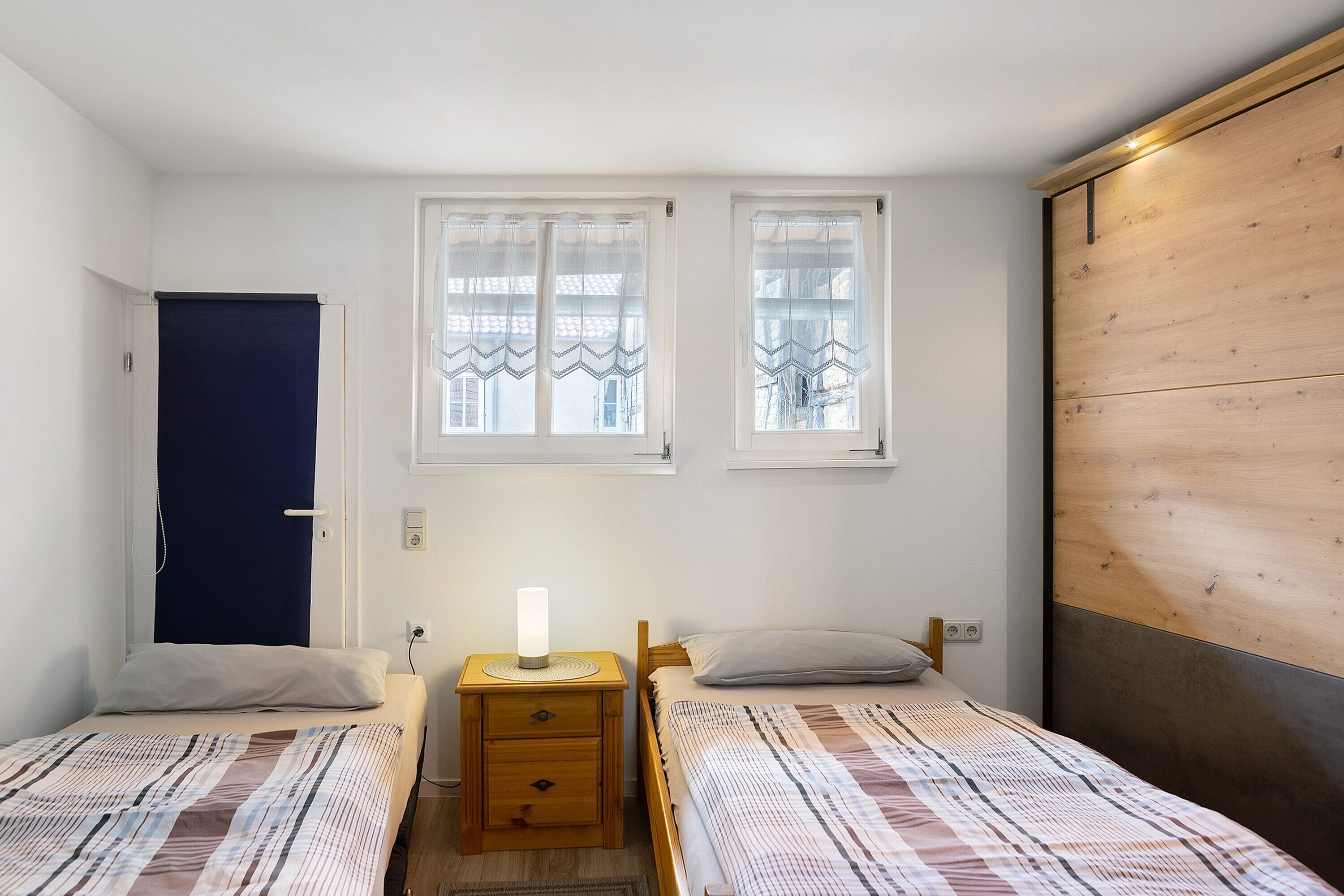 A bedroom with two beds, a nightstand with a lamp, a wooden wardrobe, and two windows with curtains.