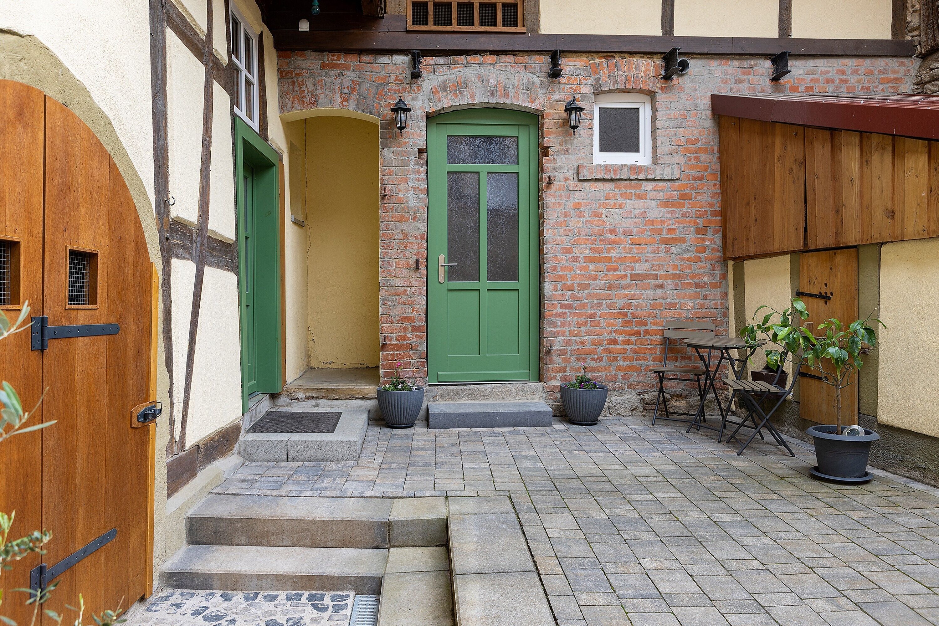 A courtyard with a green door, a wooden door, and a brick wall. There are steps leading to the door, a table with chairs, and two potted plants.