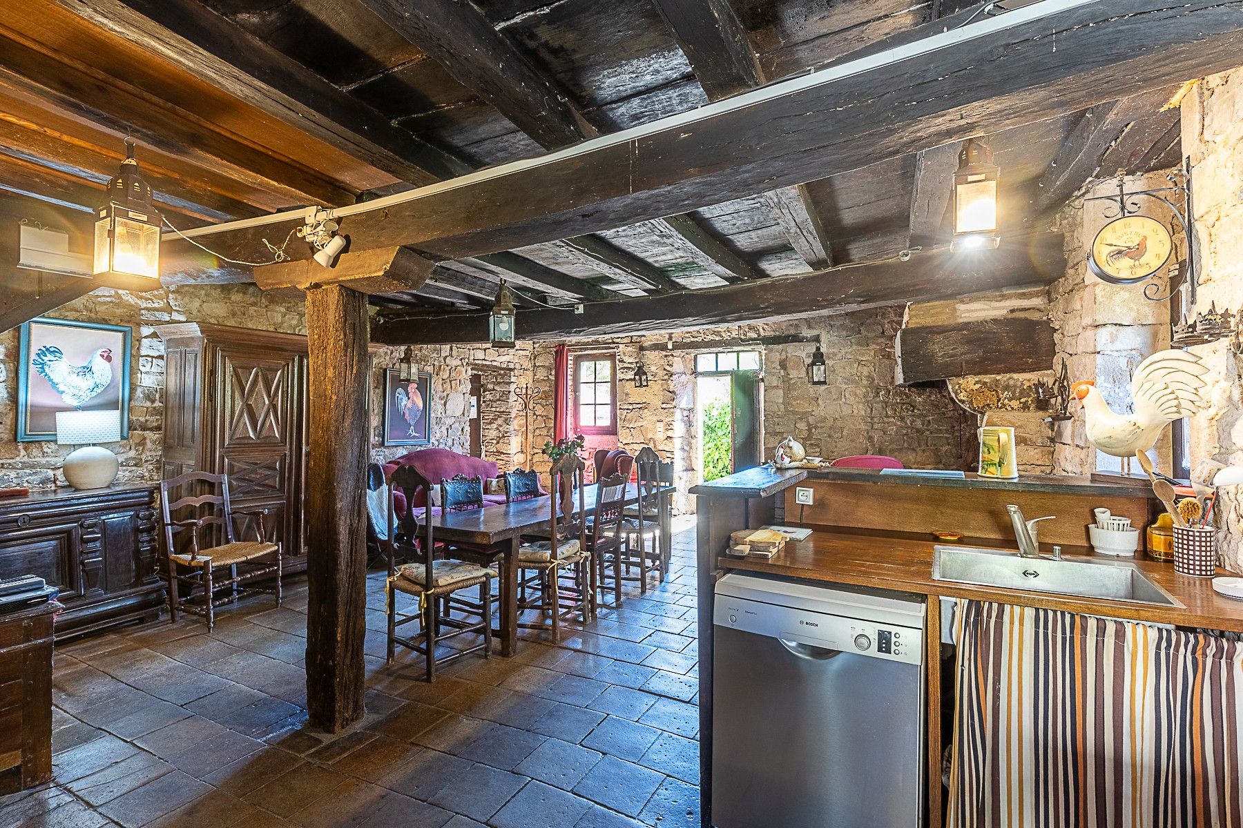 A rustic interior with exposed wooden beams, stone walls, a wooden dining table with chairs, a bar area, and a Bosch dishwasher.