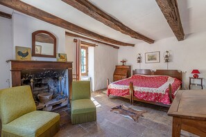 Een klassieke kamer met een open haard, een bed met een bloemenprint, een groene fauteuil en een houten tafel.