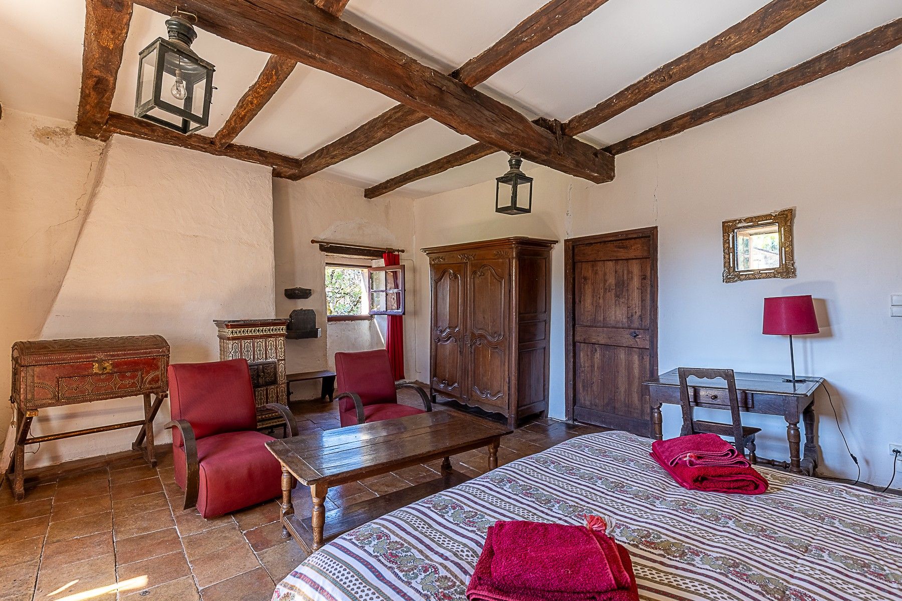 A rustic room with exposed wooden beams, a wooden wardrobe, a red upholstered chair, a wooden table, and a patterned bedspread.