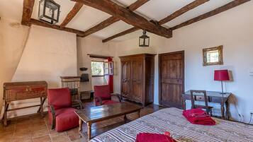 A rustic room with exposed wooden beams, a wooden wardrobe, a red upholstered chair, a wooden table, and a patterned bedspread.