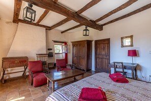 Una habitación rústica con vigas de madera a la vista, un armario de madera, un sillón tapizado de rojo, una mesa de madera y una colcha con estampado.