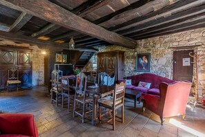 Ein rustikales Interieur mit freiliegenden Holzbalken, einer Steinwand, einem Holztisch mit Stühlen, einem roten Sofa und einer Treppe, die zu einer oberen Ebene führt.