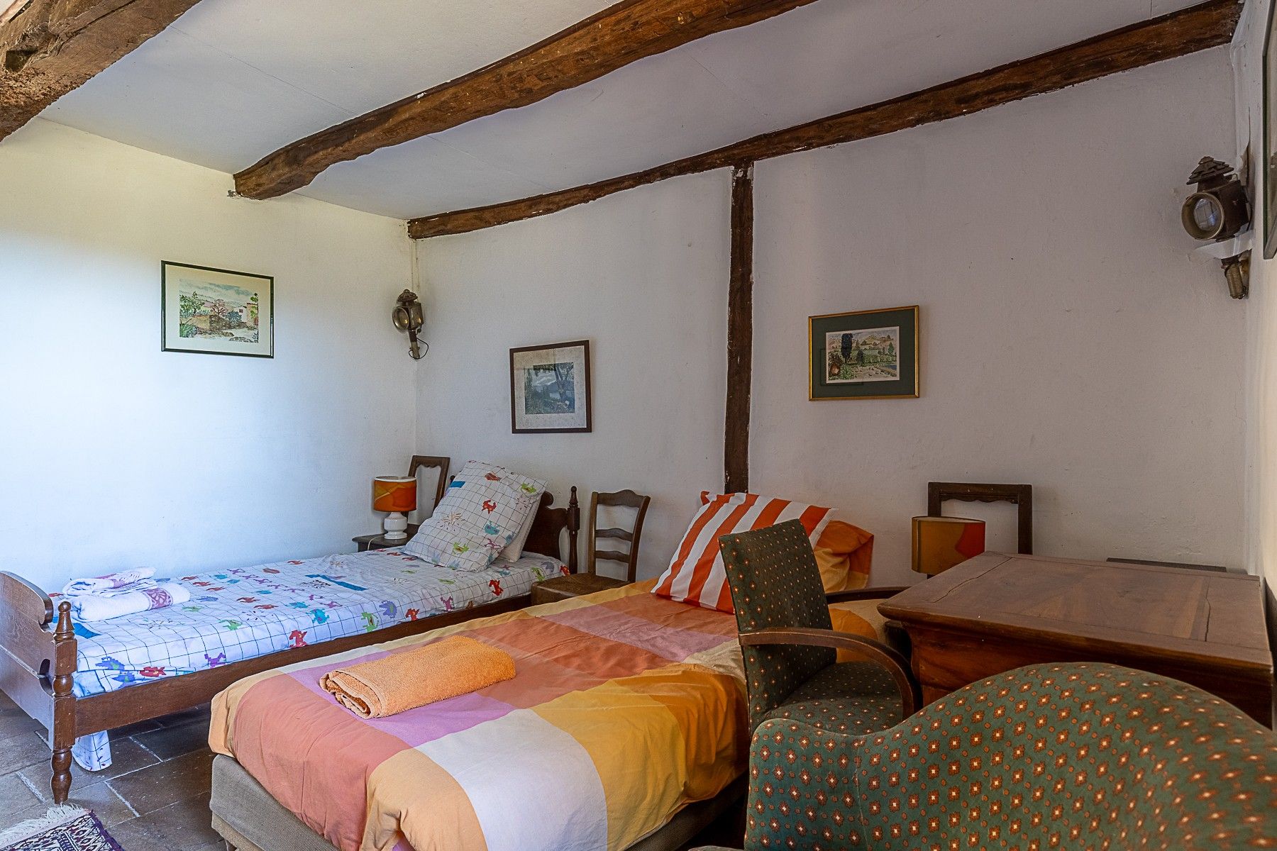 A room with two beds, a wooden chair, a table, and wall-mounted lamps.