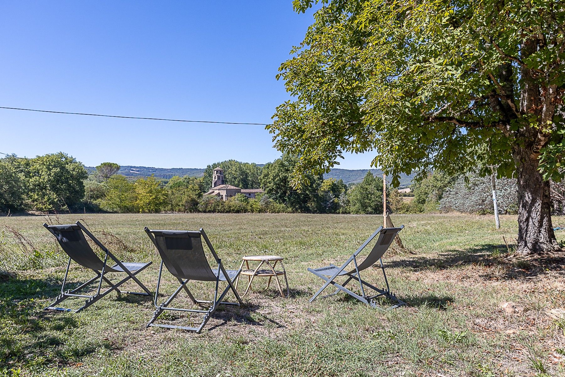 An outdoor seating area with two chairs and a small table, surrounded by grass and trees, with a view of a hill and a building in the distance.