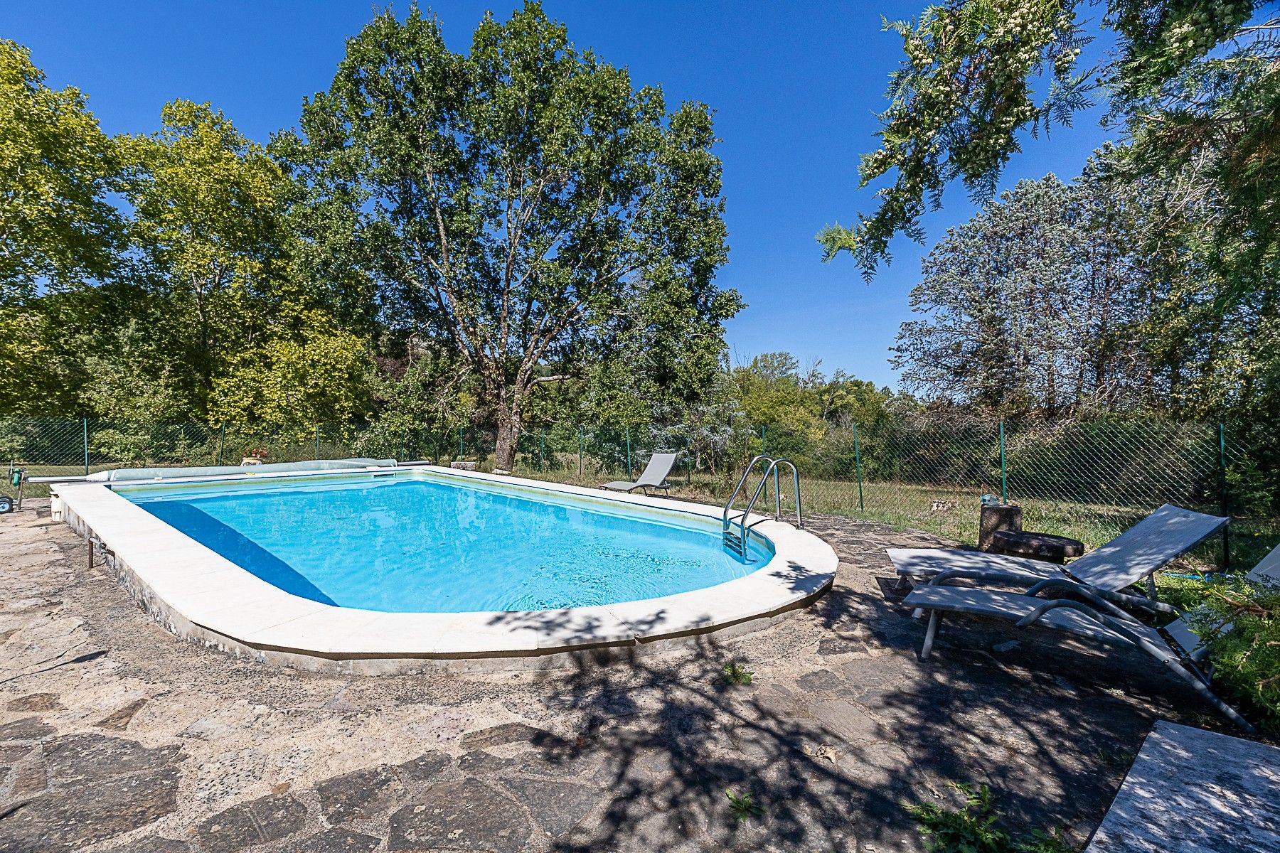 Piscina con borde de baldosas, rodeada de árboles y una valla.