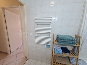 Une salle de bain avec un porte-serviettes blanc, une étagère en bois où sont pliées des serviettes, et une porte donnant sur une autre pièce.