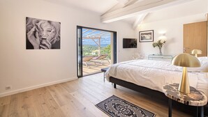 Un dormitorio con cama, mesita de noche, lámpara, un cuadro y vistas a un espacio exterior con sillas a través de una puerta de vidrio.