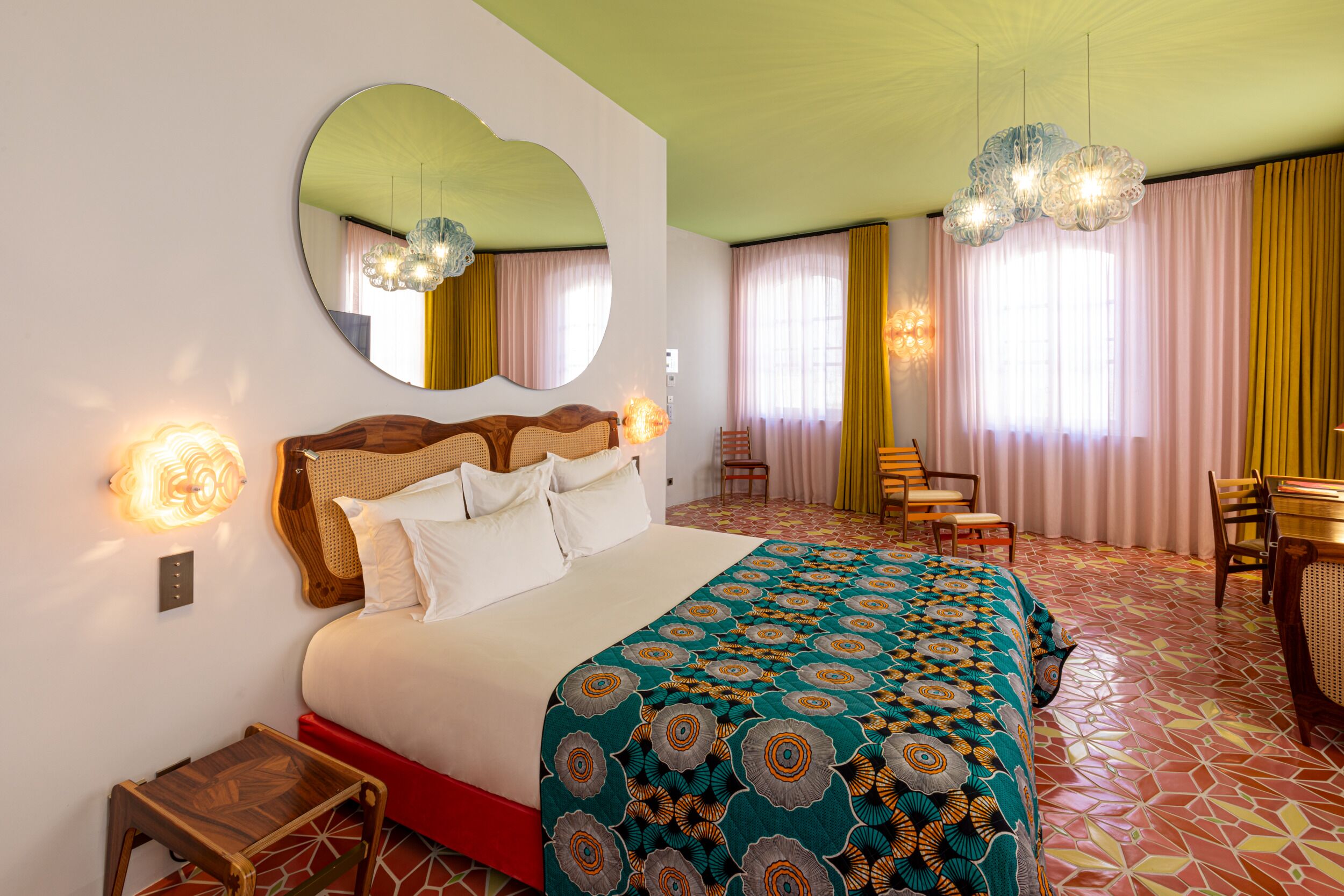 A hotel room with a large bed, a round mirror, a wooden headboard, a small table, a chair, a desk, and a patterned rug.