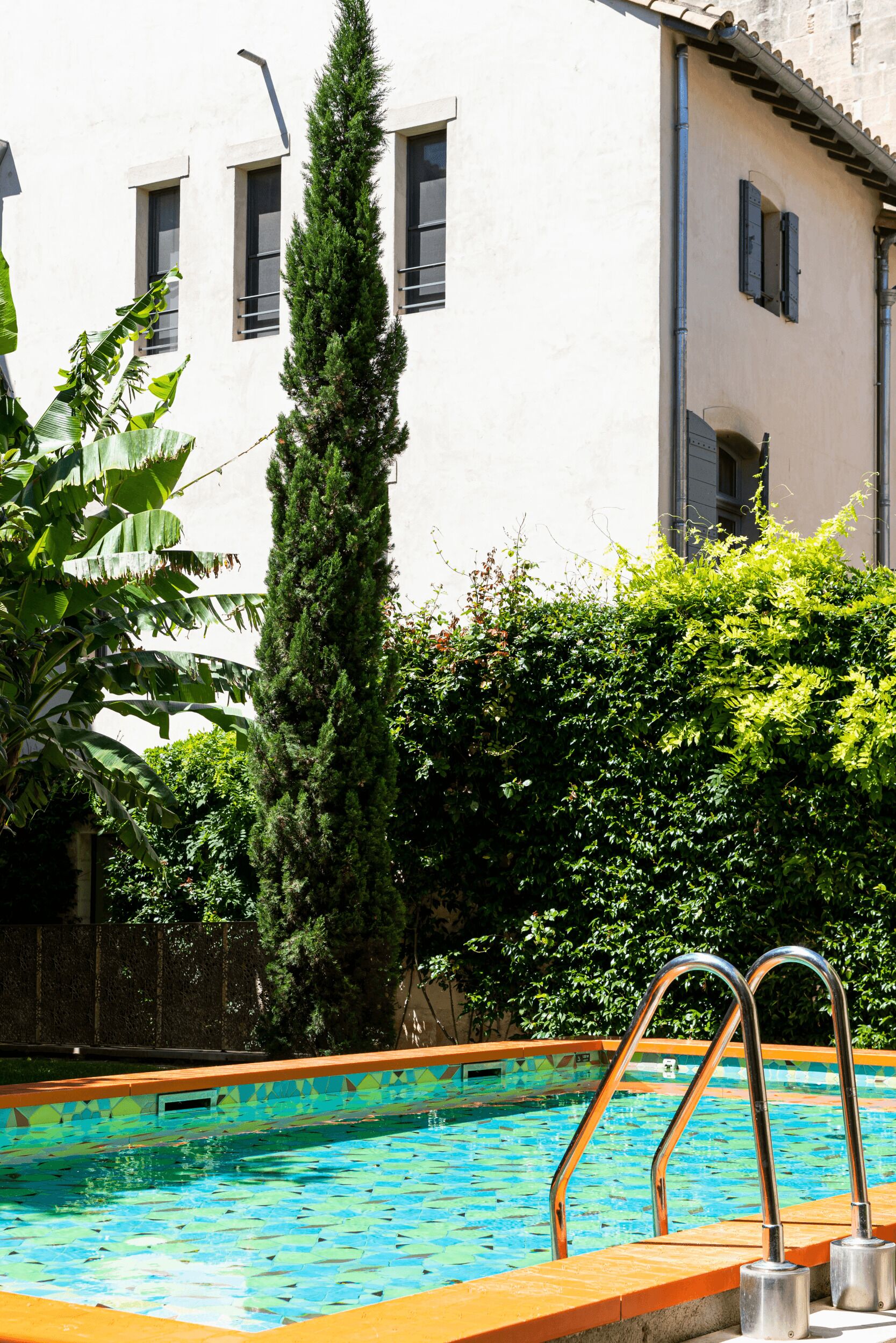 Una piscina con borde de azulejos, rodeada de follaje y un edificio con ventanas.