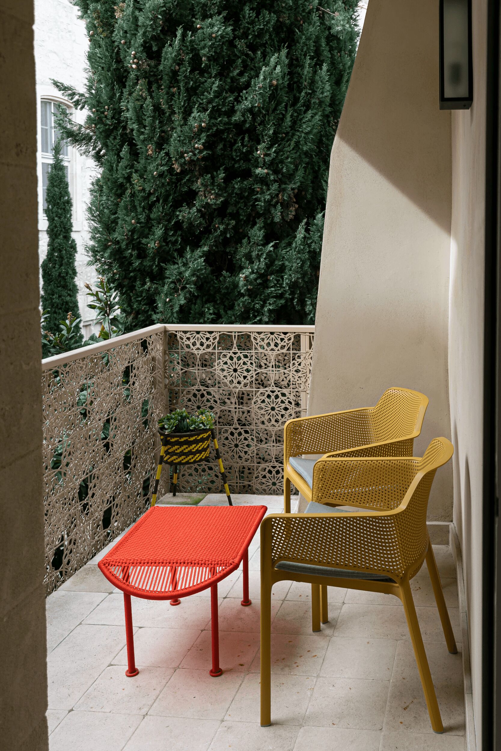 Varanda com mesa vermelha e duas cadeiras amarelas, planta em vaso e parede decorativa.