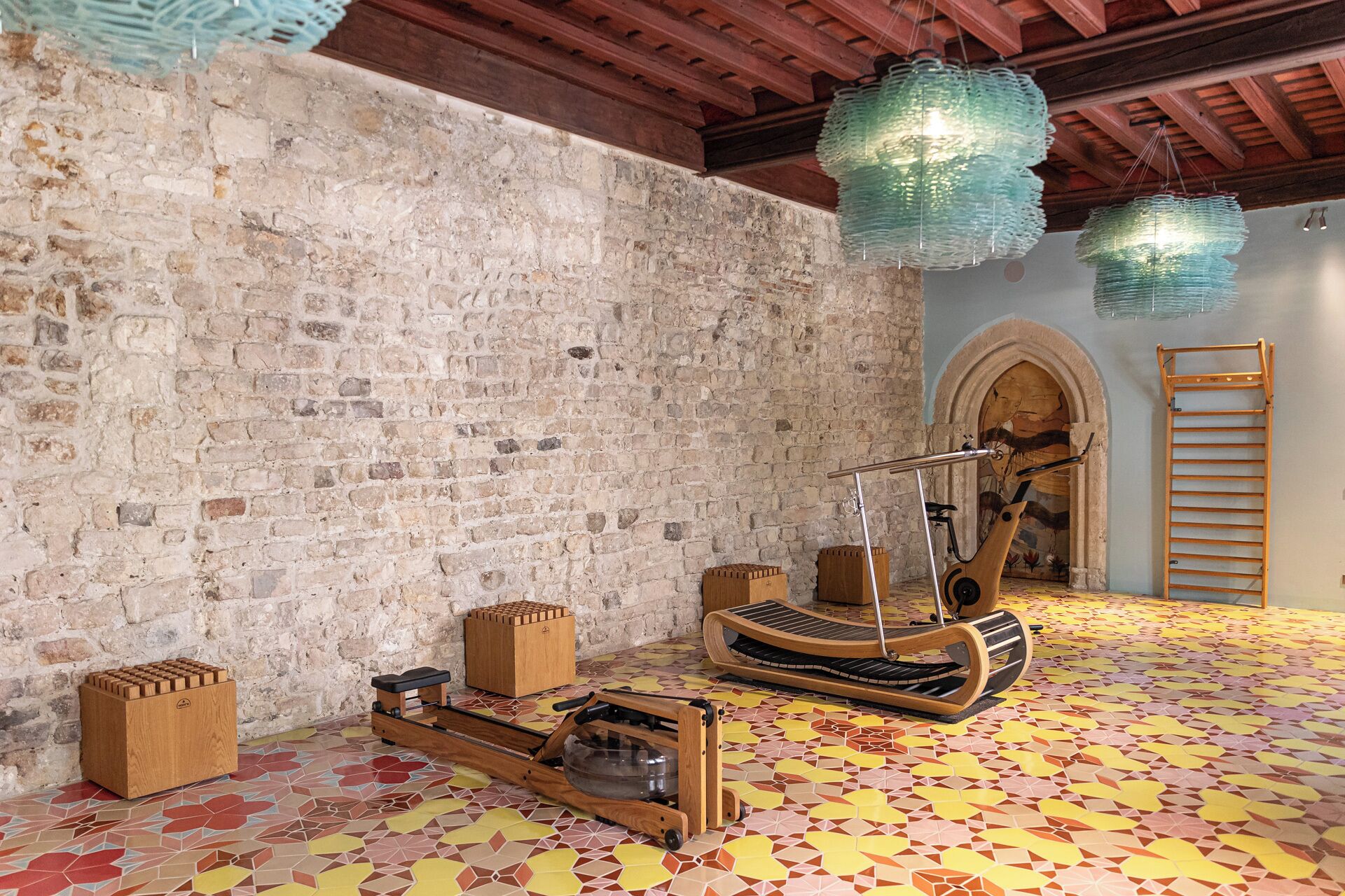 Una habitación con pared de ladrillo, suelo de madera con diseño de mosaico geométrico, máquina de ejercicio de madera y lámpara colgante.