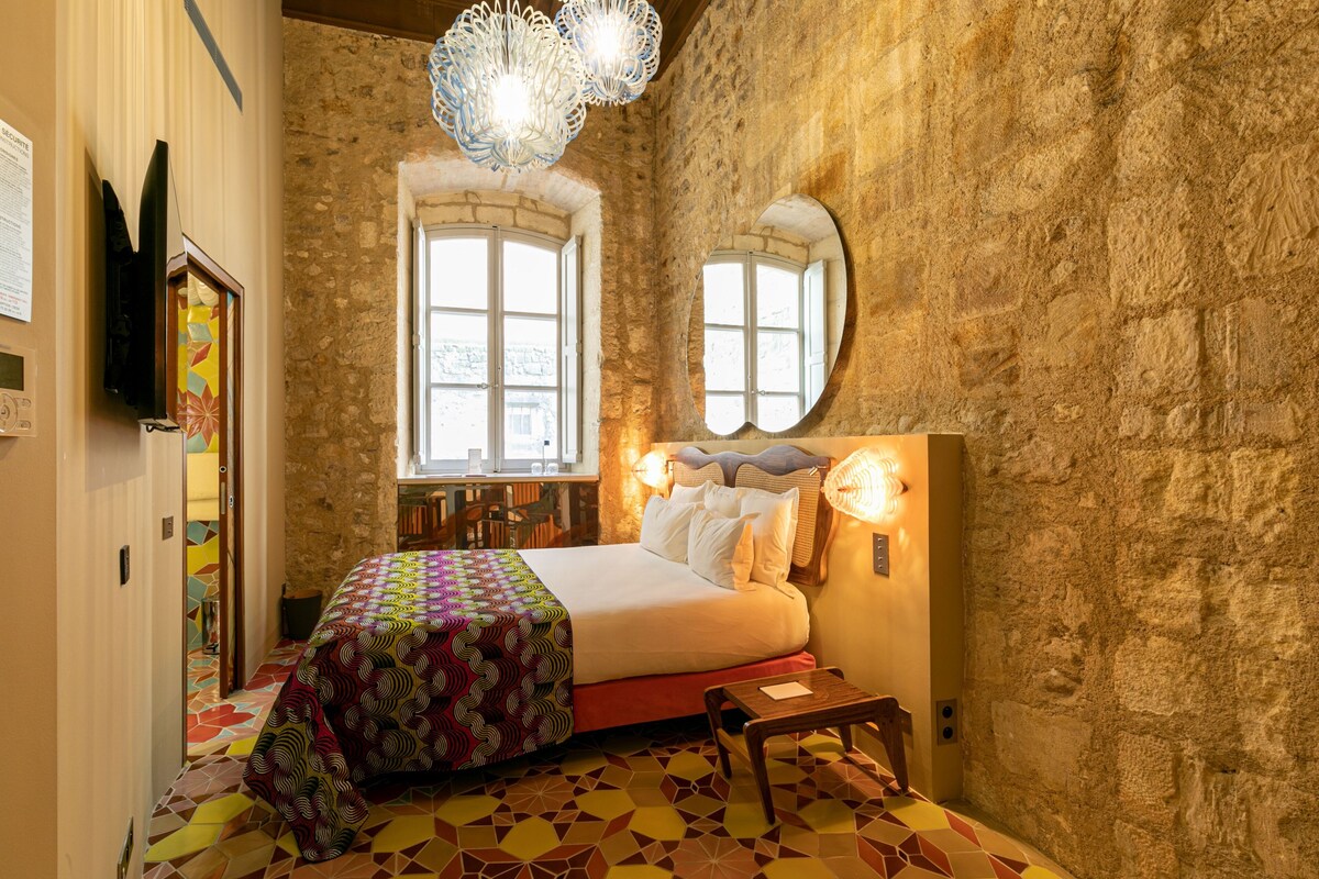 A room with a bed, a mirror, a window, and a chandelier.