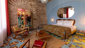 A room with a bed, a wooden table, a chair, a mirror, and a colorful floor. - L'Arlatan (Arles)