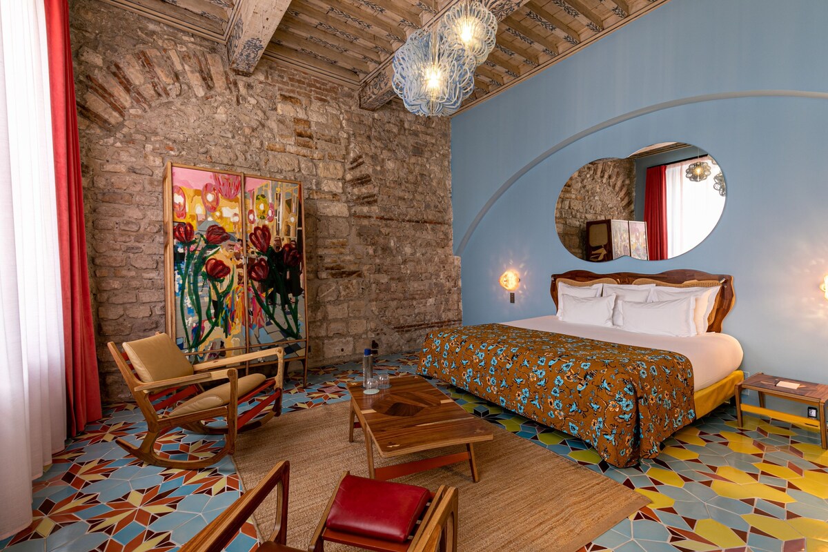 A room with a bed, a wooden table, a chair, a mirror, and a colorful floor.