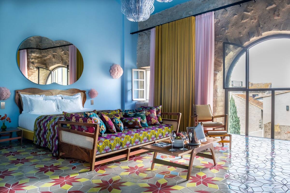 Quarto com cama, sofá, cadeiras, uma janela ampla e vista para edifícios.