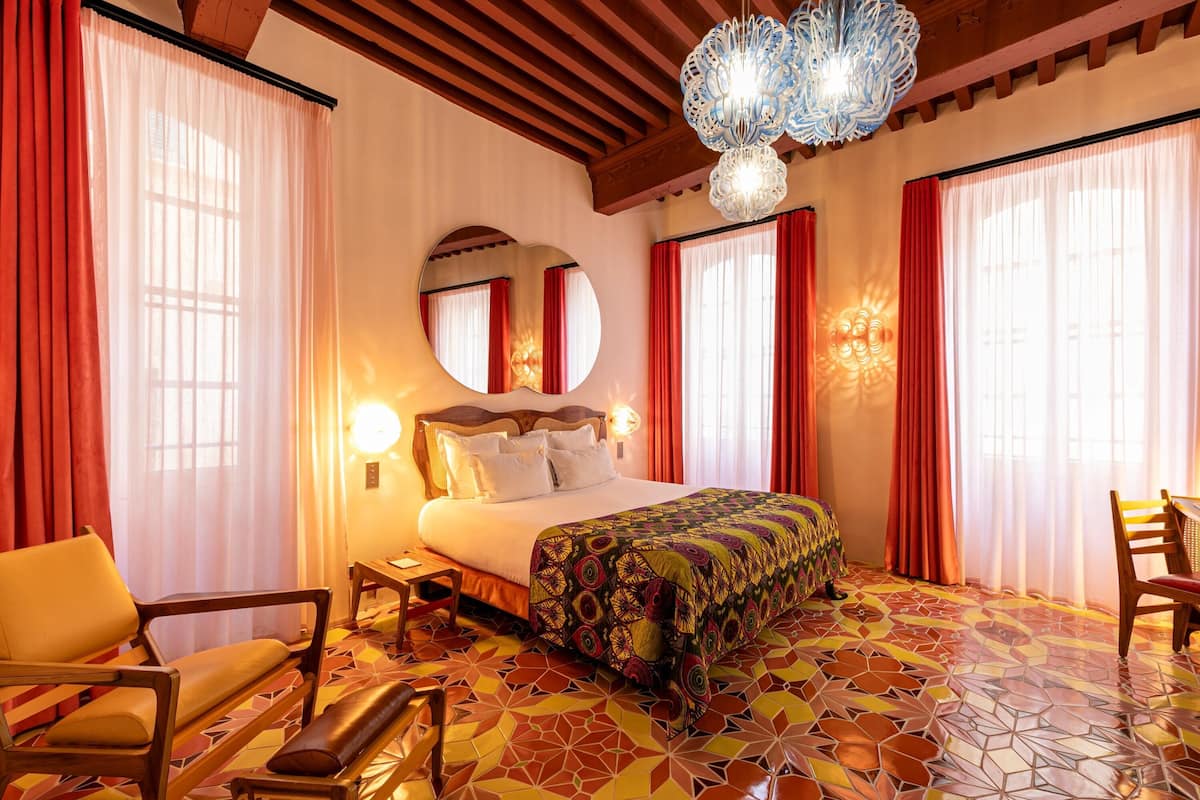 Quarto de hotel com cama, poltrona, mesa, espelho e lustre.