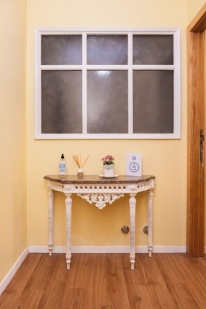 Ein Zimmer mit Holzboden, einem Fenster mit weißem Rahmen, einem kleinen Tisch mit einer Flasche, Schilferdiffusoren, einer Vase mit Blumen und einem gerahmten Zertifikat.