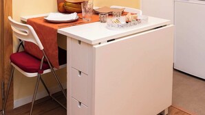 Una kitchenette compacta con cubierta blanca, una mesita con mantel rojo y una silla con cojín rojo.