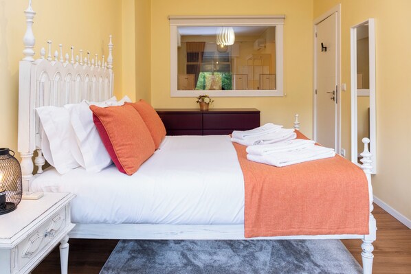 Un lit bien fait, avec une literie blanche et orange, une table de chevet avec une lampe, et une fenêtre donnant sur l’extérieur.