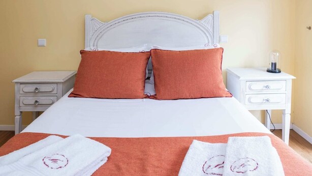 Una cama bien tendida con sábanas blancas y dos almohadas anaranjadas, flanqueada por dos mesitas de noche con lámparas, en una habitación con paredes amarillas.