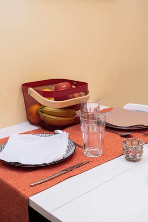 Une table recouverte d’une nappe rouge, un verre transparent, une fourchette en métal, une serviette, un verre décoratif et un panier rempli de fruits.