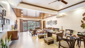 A spacious living area with a ceiling fan, large windows, and a dining area with wooden furniture. - ELIVAAS Lakeview Manor 3-BHK Luxurious (Bhimtal)