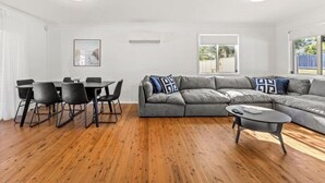 A modern living room with a grey sectional sofa, a dining table with chairs, a small coffee table, and a large window providing natural light. - Poppi's Paradise - Jervis Bay Escape | Quiet, Modern & Perfect for Families (Sanctuary Point)