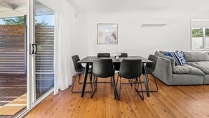 A modern living space with a dining area, a grey sofa, and a large window. - Poppi's Paradise - Jervis Bay Escape | Quiet, Modern & Perfect for Families (Sanctuary Point)