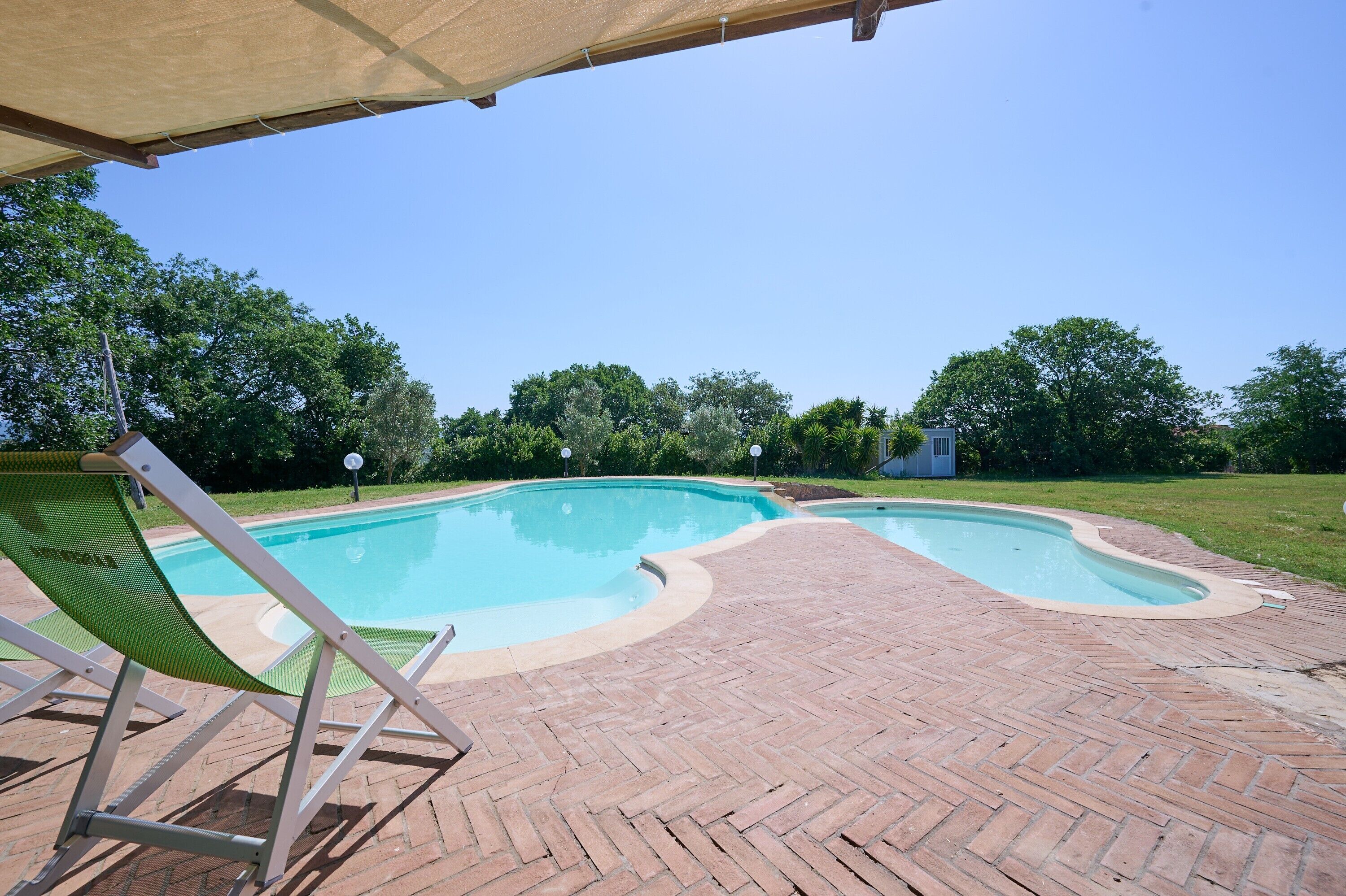 Una piscina con patio de ladrillo, una silla reclinable verde y una zona cubierta.