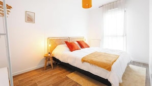 Quarto com cama, mesa de cabeceira, abajur, janela com cortinas e piso de madeira.