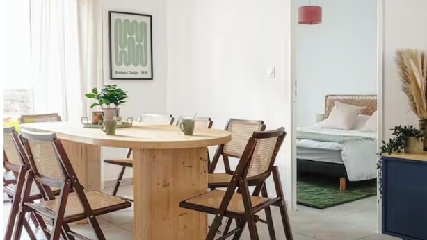 Uma sala de jantar com uma mesa de madeira redonda, quatro cadeiras, uma planta em vaso e um quadro na parede.