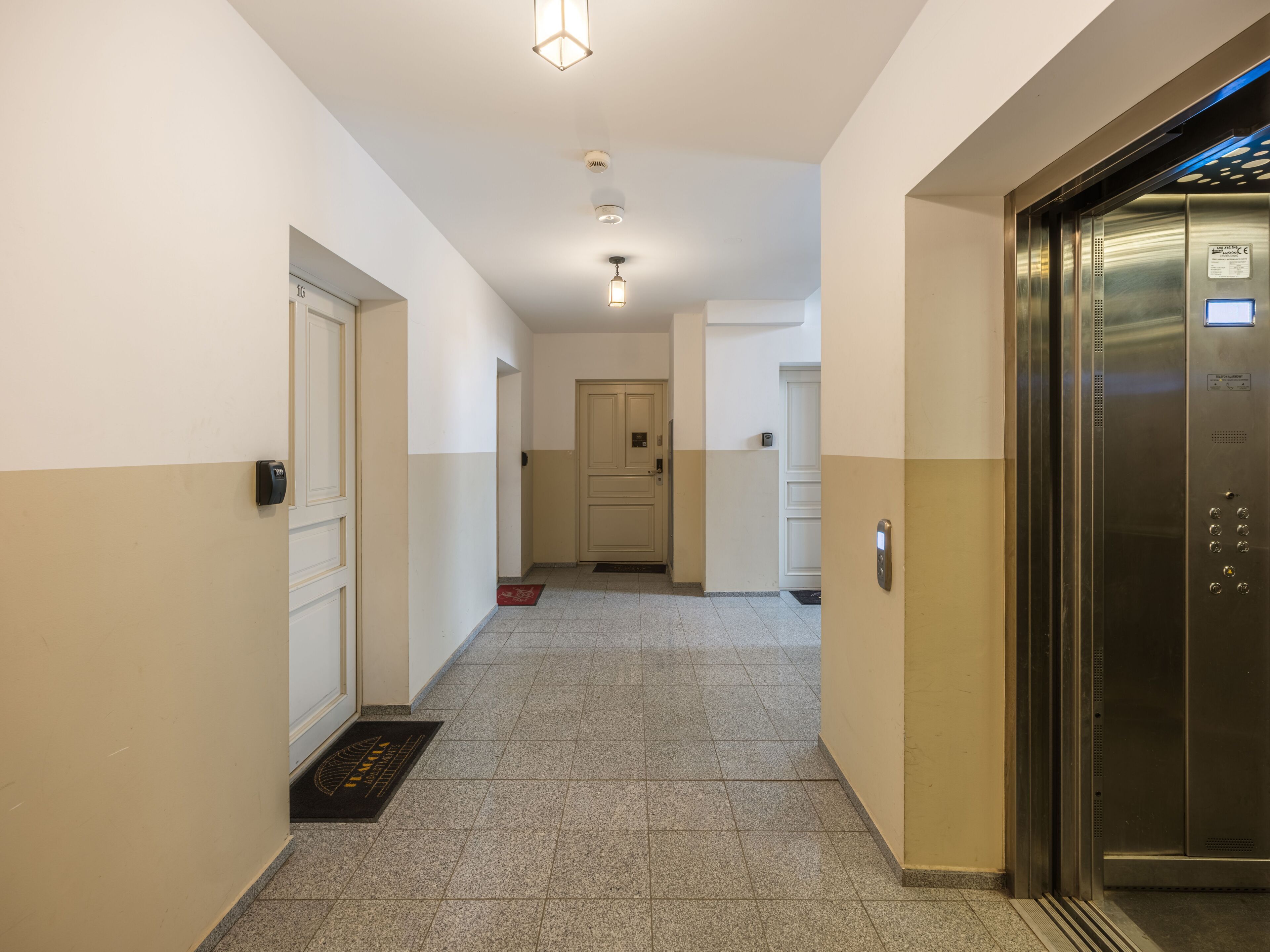 Un pasillo con una puerta a la izquierda, un felpudo y un ascensor a la derecha.