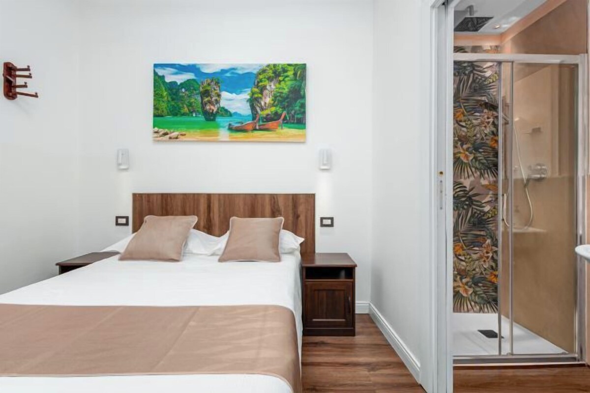 A hotel room with a bed, a nightstand, a painting, and a shower area.