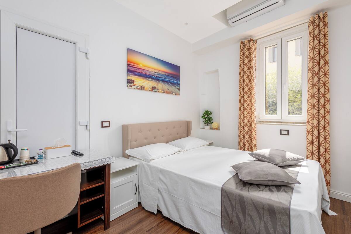 A bedroom with a bed, a desk, a chair, a window with curtains, and a painting on the wall.