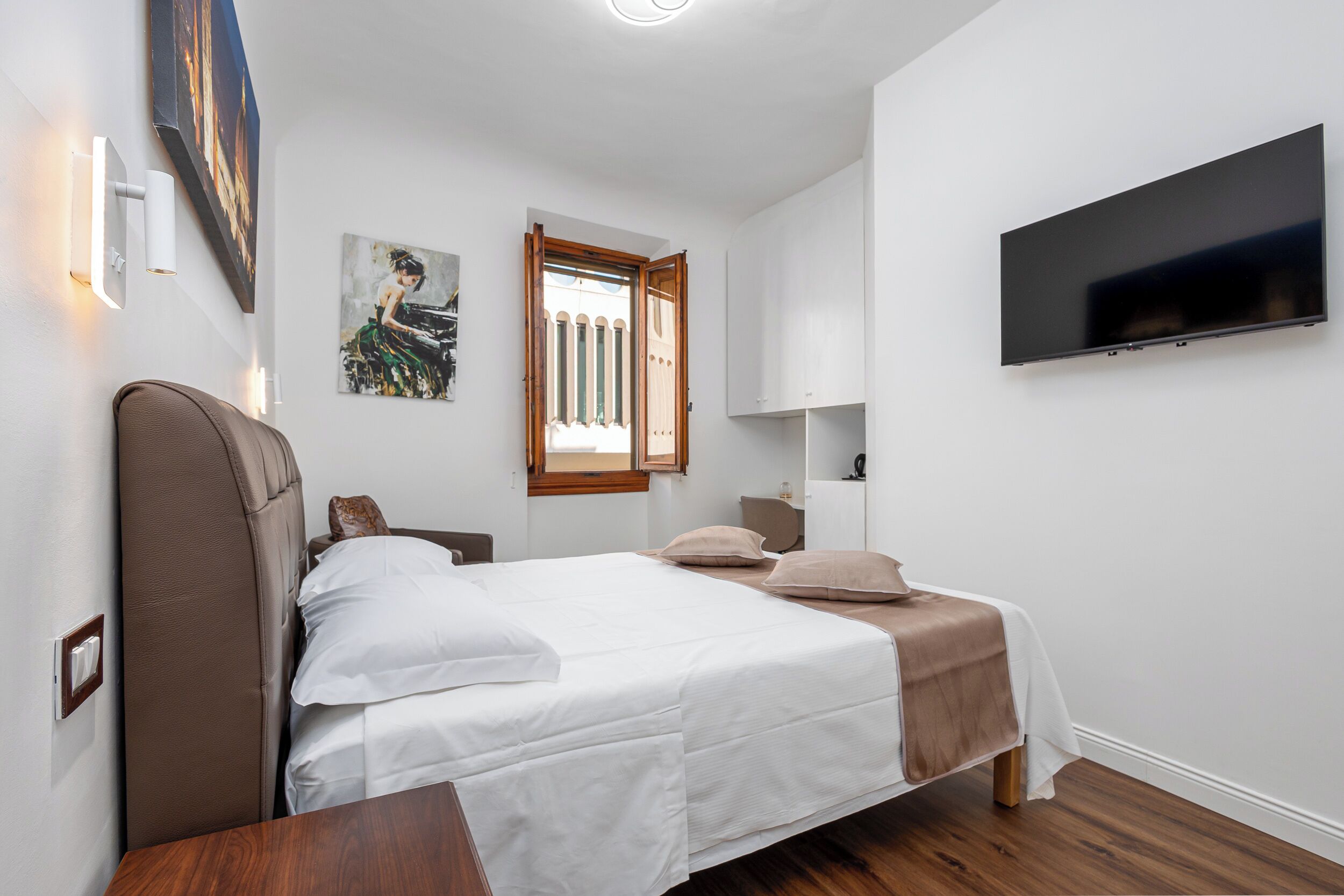 A bedroom with a bed, a television, a painting, and a window.