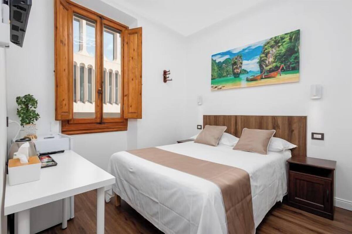 A bedroom with a bed, a desk, a window with wooden shutters, a painting on the wall, and a small plant.