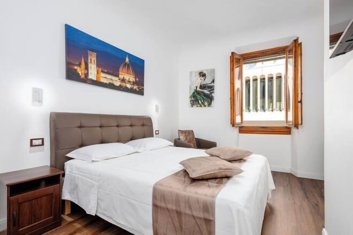 A bedroom with a large bed, a nightstand, a wooden cabinet, a window with a view, and two paintings on the wall.