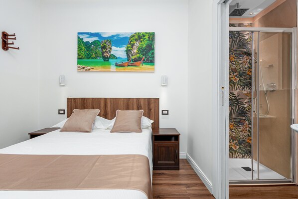 A bedroom with a bed, a nightstand, a wooden headboard, a painting of a tropical scene, and a shower with a glass door.