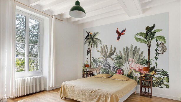 Une chambre avec un grand lit, une fenêtre avec des rideaux, un radiateur et une fresque murale sur le thème des tropiques.