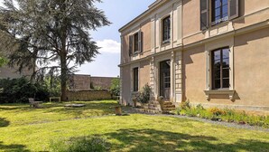 Un bâtiment à deux étages avec un jardin devant, un banc et un arbre.
