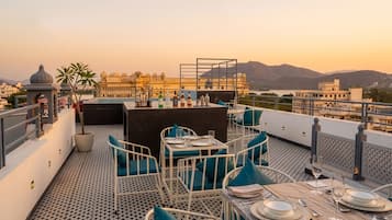 Terraço no telhado com mesas preparadas para refeições, vista para um palácio e montanhas ao fundo.