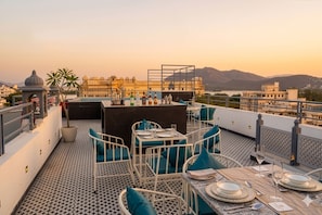 A rooftop terrace with tables set for dining, a view of a palace, and a mountainous backdrop. - UDAISA - The Luxury Art (Udaipur)