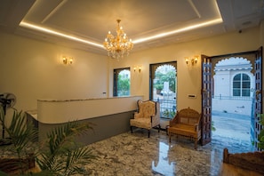 Un área de recepción con suelo de mármol, una araña de cristal, un mostrador de recepción, un sillón tapizado con flores y un banco de madera.